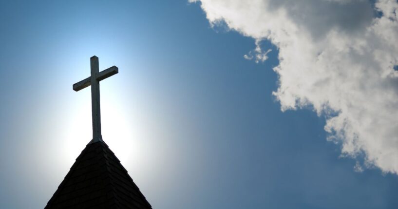 A wood cross sits on top of an old church steeple in front of a blue sky.