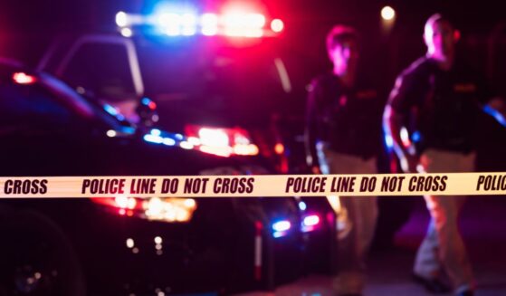 Two police officers standing in front of patrol cars, behind police tape, wearing bulletproof vests and duty belts.