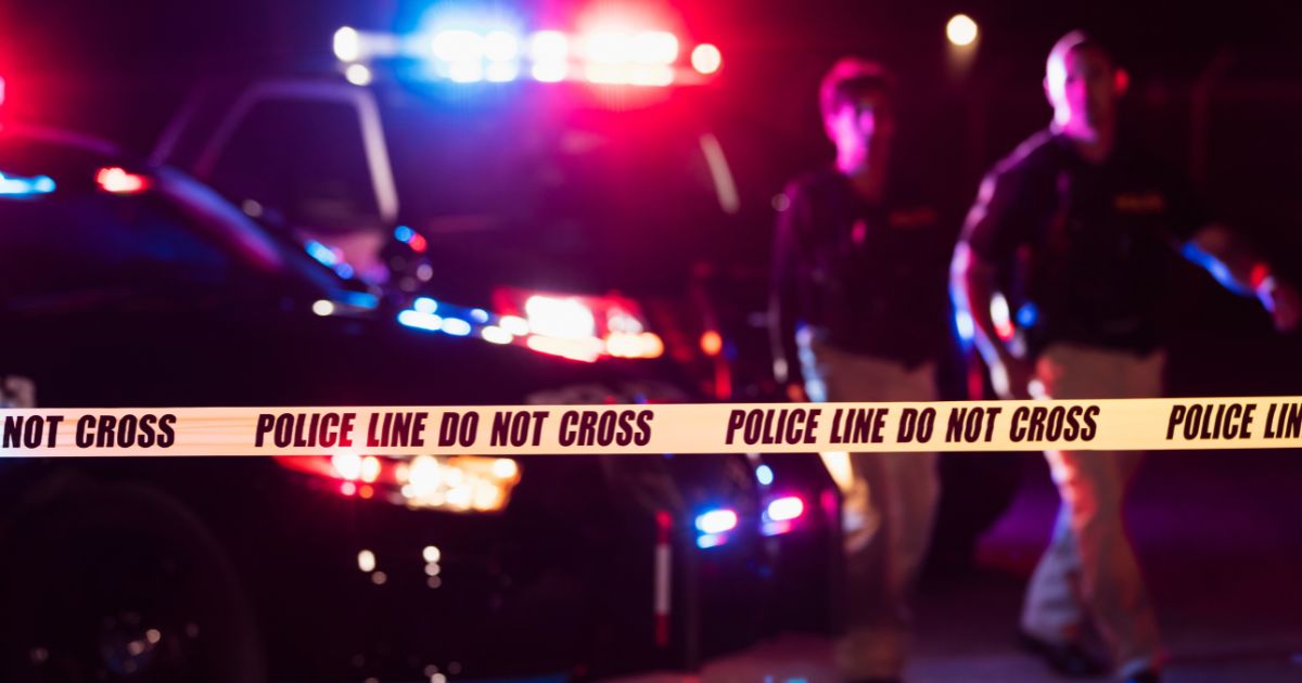 Two police officers standing in front of patrol cars, behind police tape, wearing bulletproof vests and duty belts.