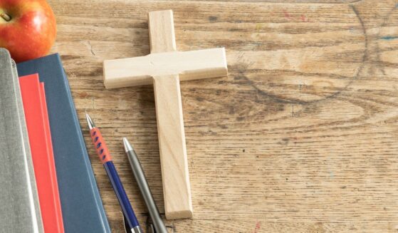 A wooden cross sits on a vintage wooden desk next to some pencils and a stack of notebooks.