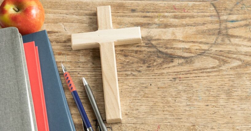 A wooden cross sits on a vintage wooden desk next to some pencils and a stack of notebooks.