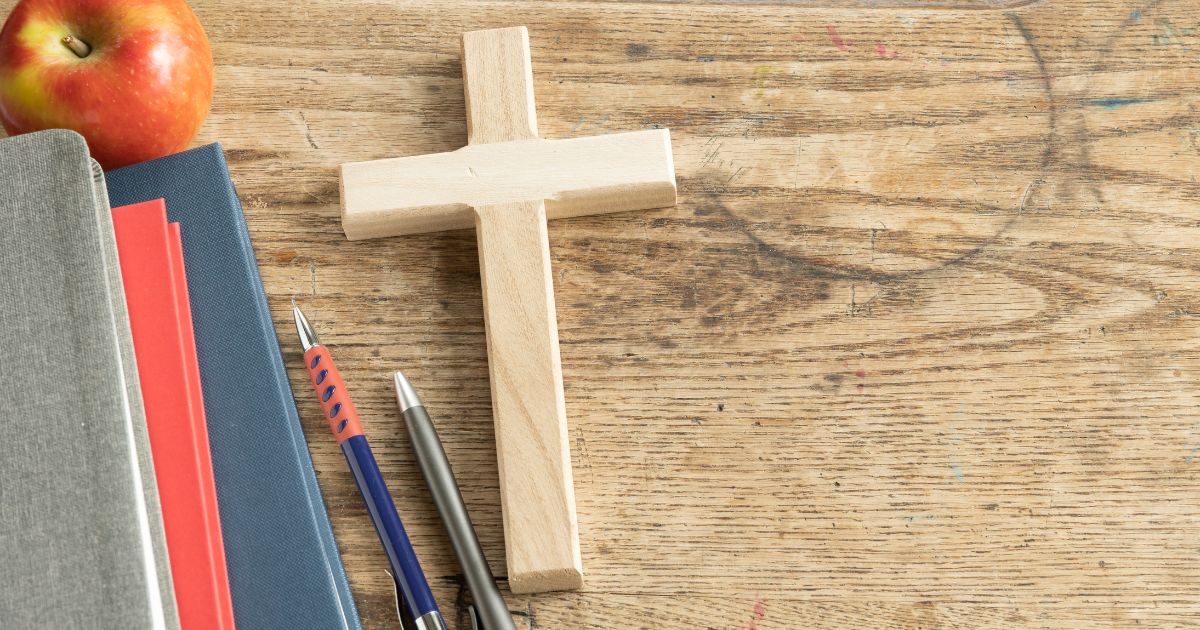A wooden cross sits on a vintage wooden desk next to some pencils and a stack of notebooks.