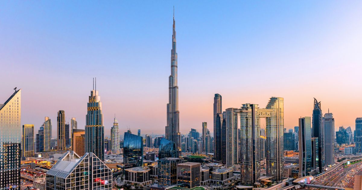 An aerial cityscape of Dubai at sunset with modern skyscrapers, illuminated highways, busy traffic, and dense urban development.