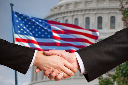 Two men shake hands as the American flag waves behind the United States Capitol rotunda.