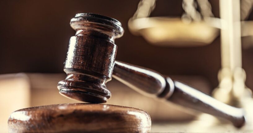 A gavel lays on a wooden block next to a balancing scale in a dark room.