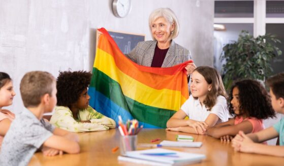 In a classroom, a woman holds up a rainbow colored flag.