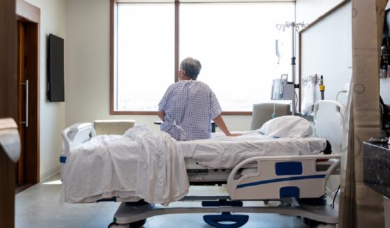 A photo illustration depicts an older woman seated on a hospital bed alone in her room.
