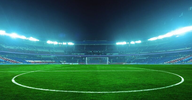 An illuminated soccer stadium at night with a green field and empty stands, ready for an exciting match under bright blue floodlights.