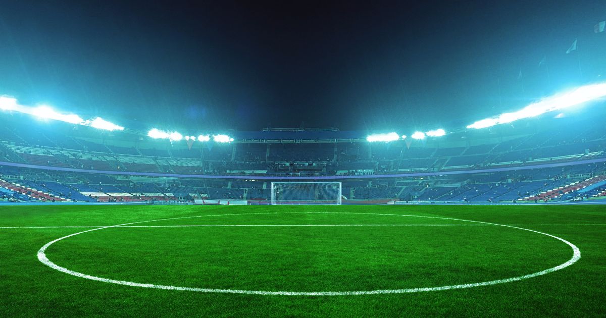 An illuminated soccer stadium at night with a green field and empty stands, ready for an exciting match under bright blue floodlights.