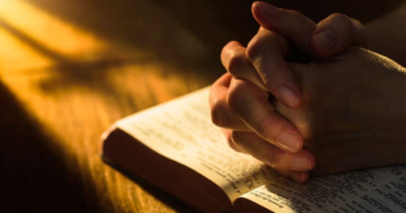 Hands clasped in a prayerful position over an open Bible.