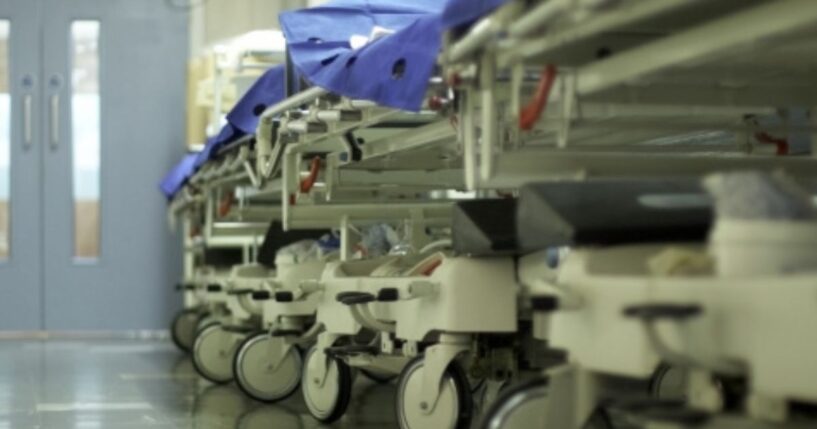 A hospital hallway is filled with gurneys and trolleys lined up on the wall.