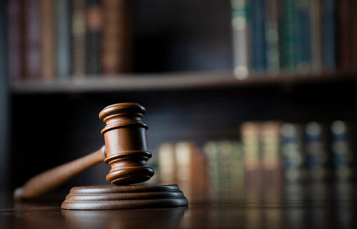 A gavel and wooden block sit on a table while book shelves sit behind them.