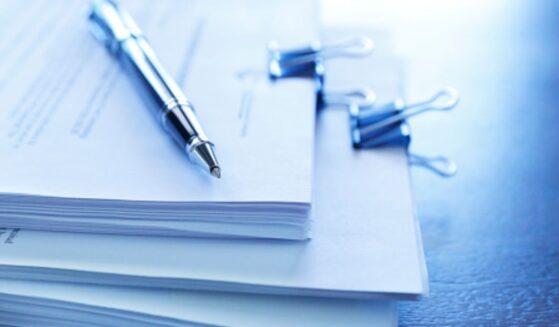 Stacks of paperwork sit on a table with a ballpoint pen topping the stack.