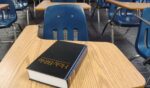 A bible sits on a school desk in a classroom.