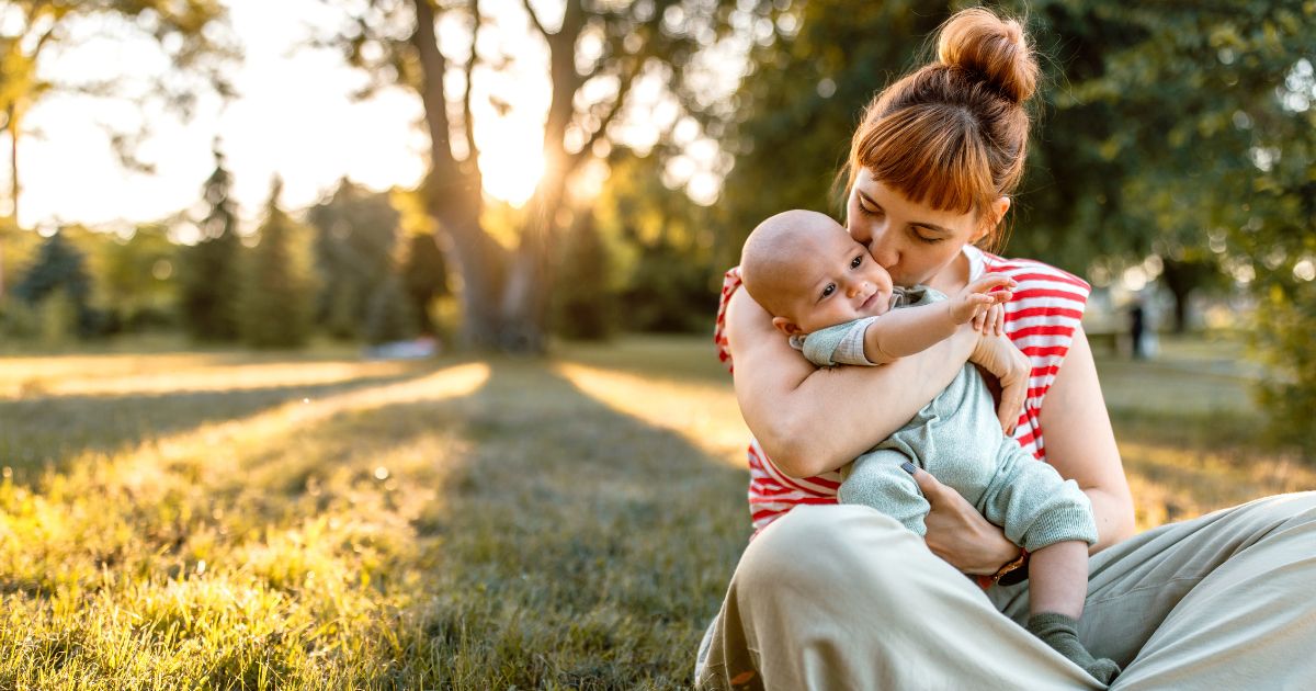 A mother and her baby embrace while sitting down in a meadow in sunset.