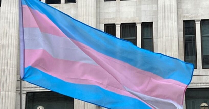 The trans flag waves outside of a building.