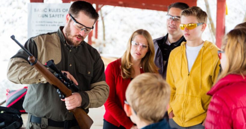A family watches a gun instructor as he teaches them about a rifle.