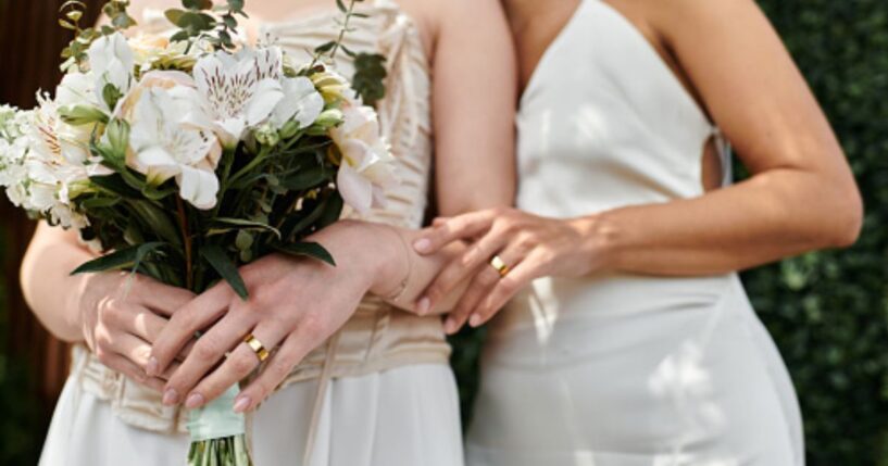 Two women dressed as brides stand close to each other as one embraces the other.