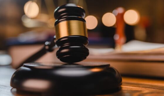 A gavel sits on a wooden block as a balancing scale and book are pictured behind.