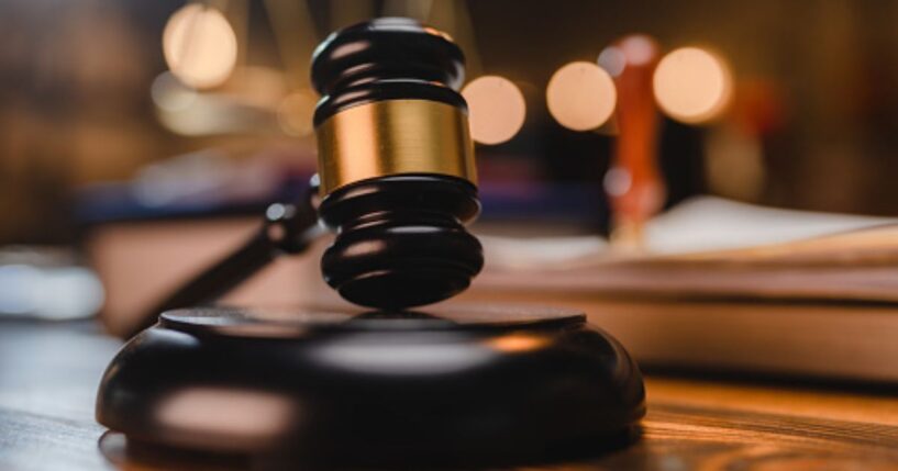 A gavel sits on a wooden block as a balancing scale and book are pictured behind.