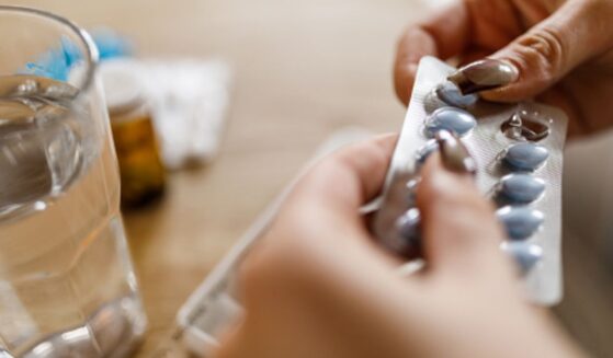 A woman opens a pill packet next to a cup of water sitting on a wooden table.