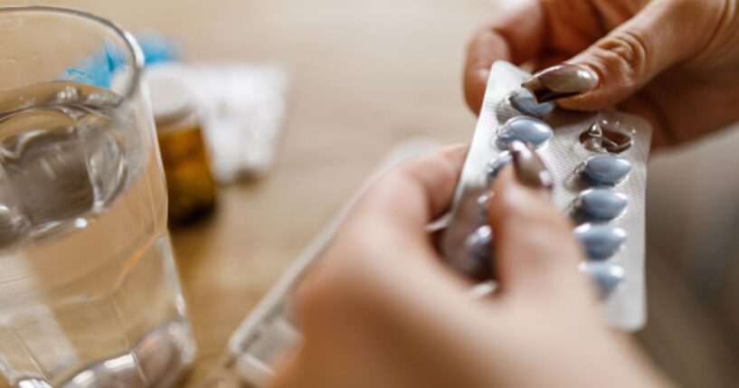 A woman opens a pill packet next to a cup of water sitting on a wooden table.