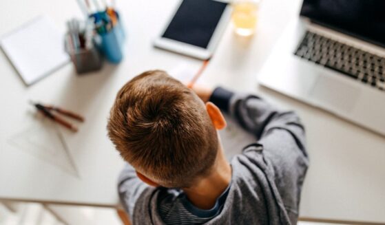 A boy does school work on a desk next to a computer, tablet and various schools supplies.