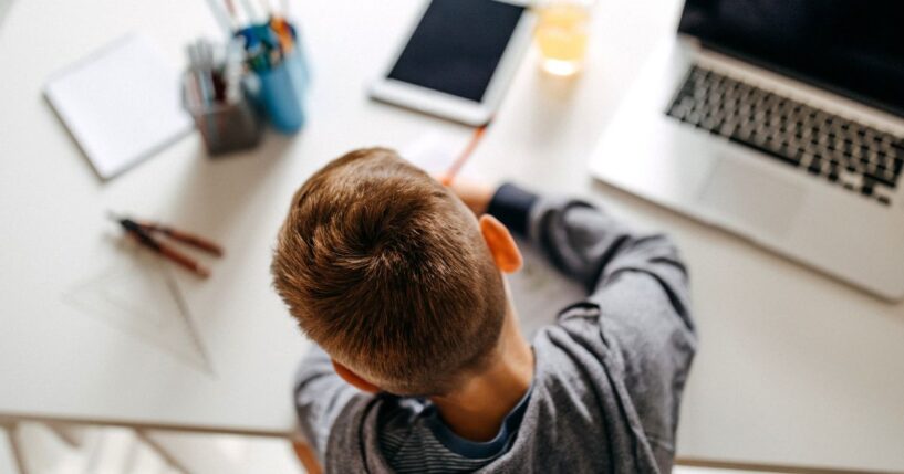 A boy does school work on a desk next to a computer, tablet and various schools supplies.