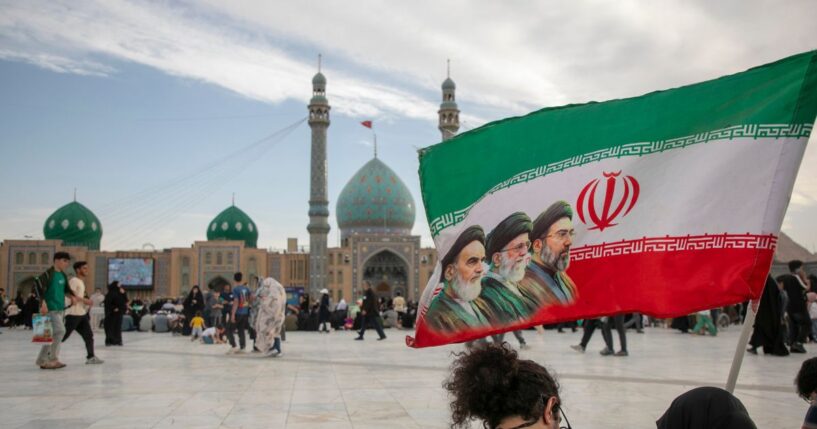 People hold a flag with Iran's late and new supreme leaders, Ayatollah Ruhollah Khomeini, Ayatollah Ali Khamenei, and Ayatollah Mojtaba Khamenei, during Eid al-Adha at the entrance of Jamkaran Mosque on May 26, 2026, in Qom, Iran.