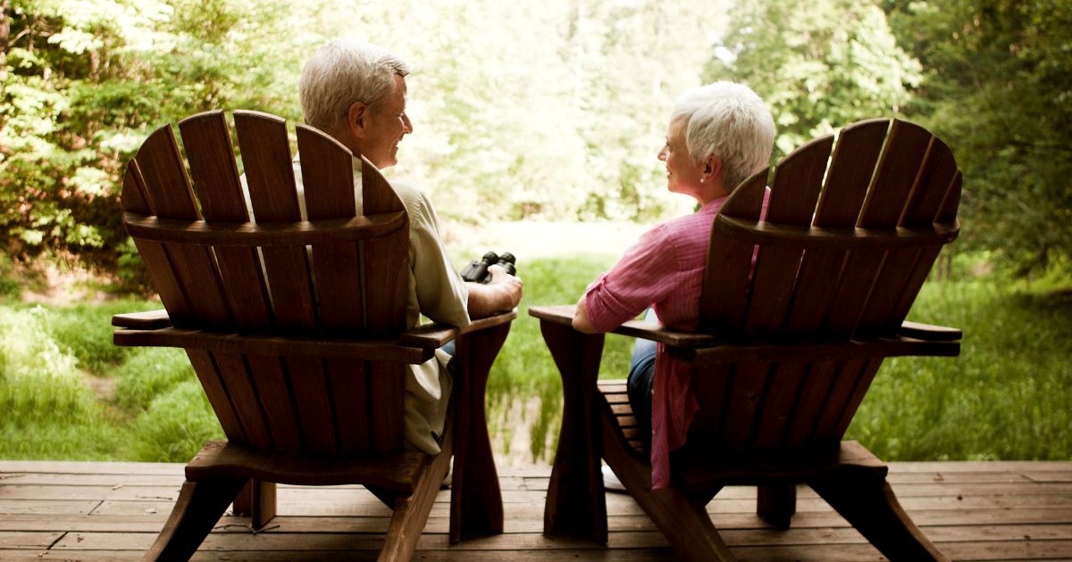 The rear view of a senior couple relaxing in Adirondack chairs on a wooden deck.