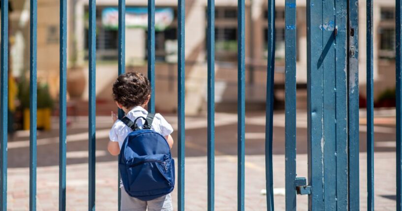 A child with a school backpack look on schoolyard grounds toward an open entrance door.