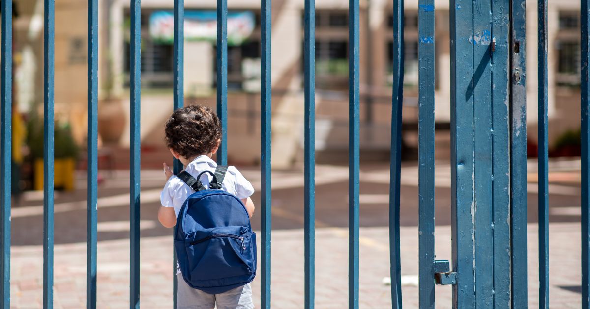 A child with a school backpack look on schoolyard grounds toward an open entrance door.