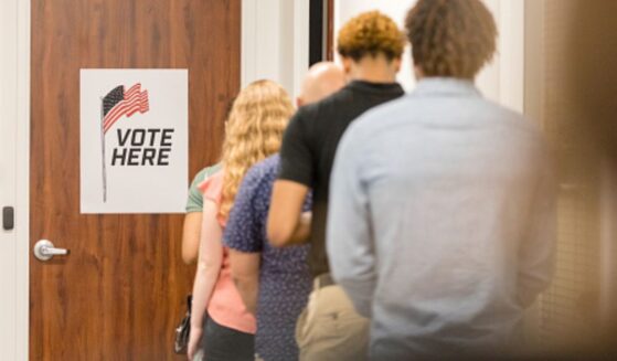 A line of multicultural citizens stand behind a door that has a sign that reads “vote here.”