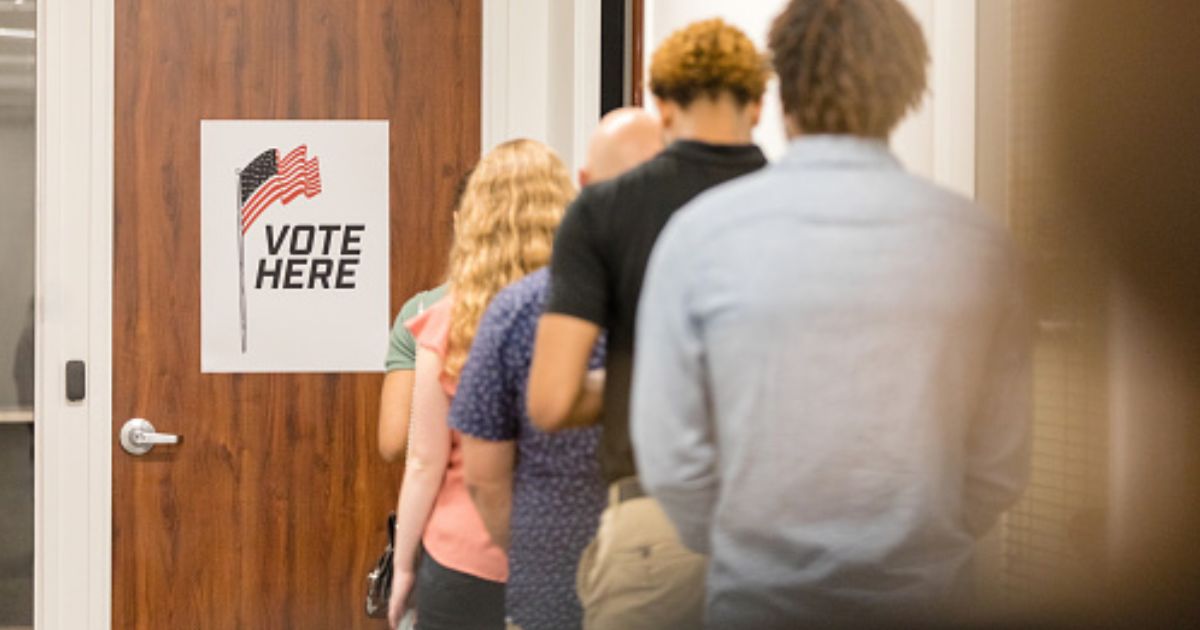 A line of multicultural citizens stand behind a door that has a sign that reads “vote here.”