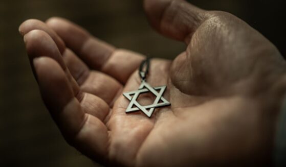 A Star of David necklace sits in a man’s palm in a dimly lit room above a wooden table.