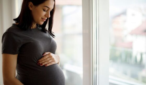 A woman rubs her stomach bulge near a window.