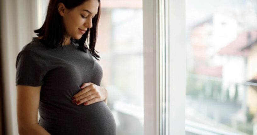 A woman rubs her stomach bulge near a window.