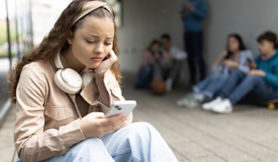 A young woman sits on a step on her phone outside a building on a bleak day ahead of a group of teens.
