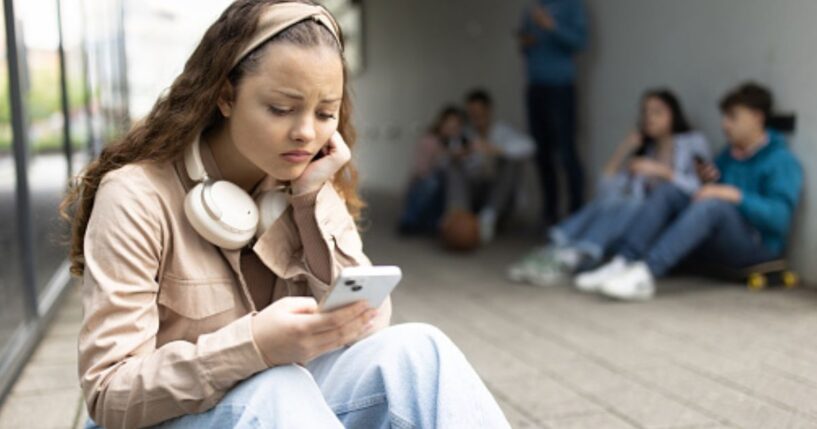 A young woman sits on a step on her phone outside a building on a bleak day ahead of a group of teens.