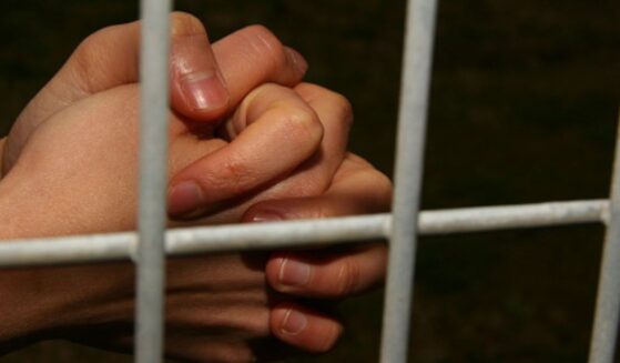 Hands are clasped behind jail bars in a dark cell.