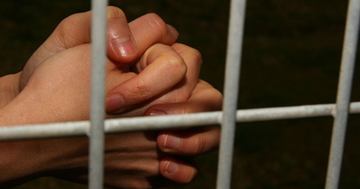 Hands are clasped behind jail bars in a dark cell.