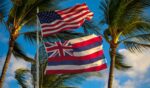 The American flag flies above the flag of Hawaii ahead of palm trees.
