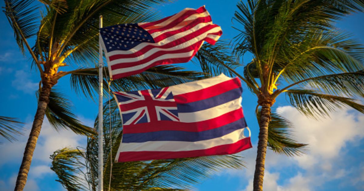 The American flag flies above the flag of Hawaii ahead of palm trees.