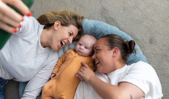 A same-sex couple of women lay on the ground with a baby as it stares up at a phone.