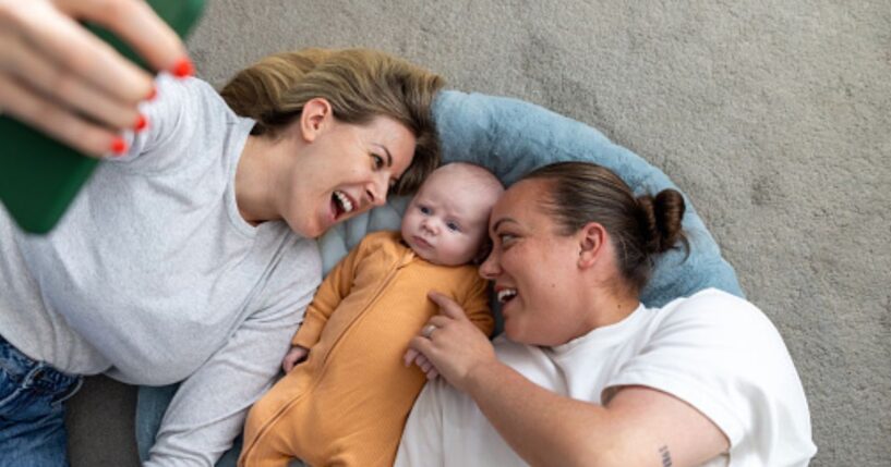 A same-sex couple of women lay on the ground with a baby as it stares up at a phone.
