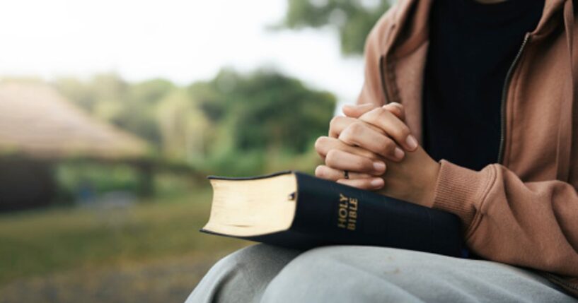 A brown woman prays with the Holy Bible in her lap outside.