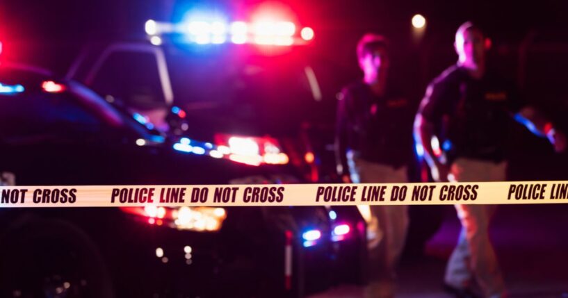 Two police officers standing in front of patrol cars, behind police tape, wearing bulletproof vests and duty belts.