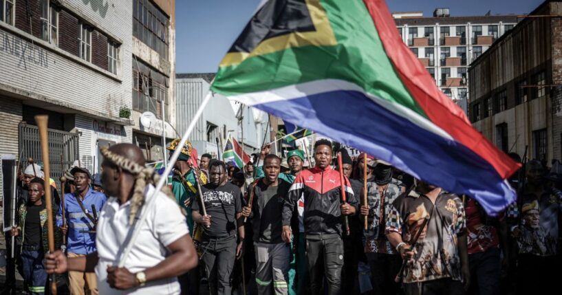 Protesters hold sticks and flags while chanting slogans as they march during a demonstration by the "March & March" and Operation Dudula movements marking an unofficial deadline set by citizen-led groups for undocumented foreign nationals to leave South Africa, in Johannesburg, on June 30, 2026.
