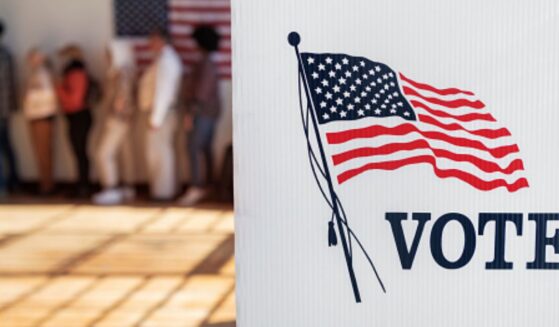 A voting booth stands in an auditorium with a line of citizens lined up behind it.