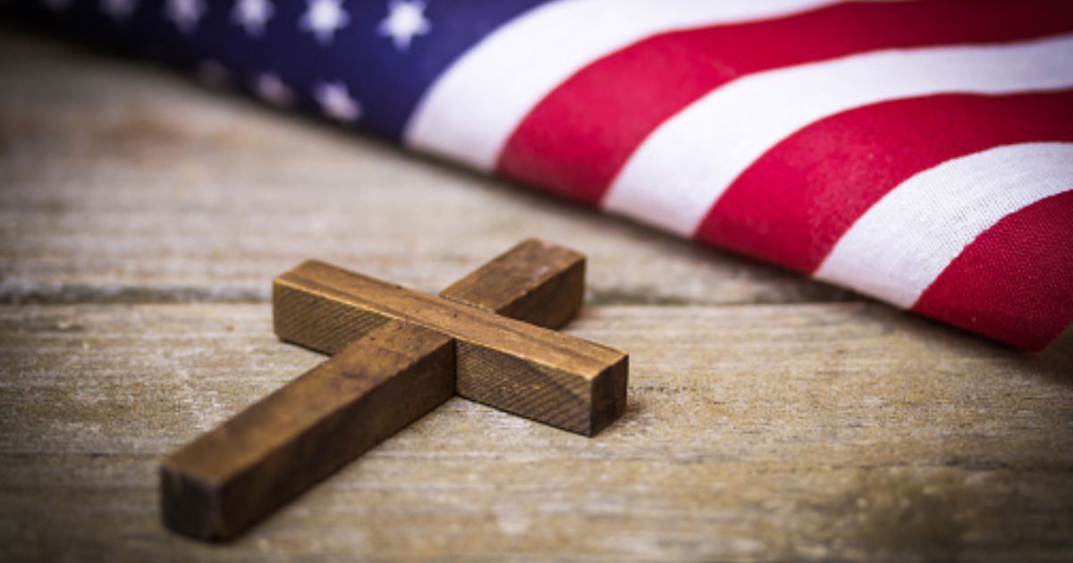 A cross and an American flag sit next to each other on a wooden table.